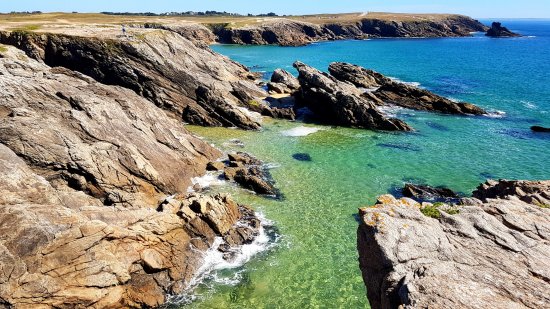 The image captures a rocky coastline where jagged cliffs meet clear turquoise waters. Gentle waves lap against the rocks, creating a tranquil scene. Lush greenery outlines the beach, leading to a distant horizon of rolling land under a bright blue sky.