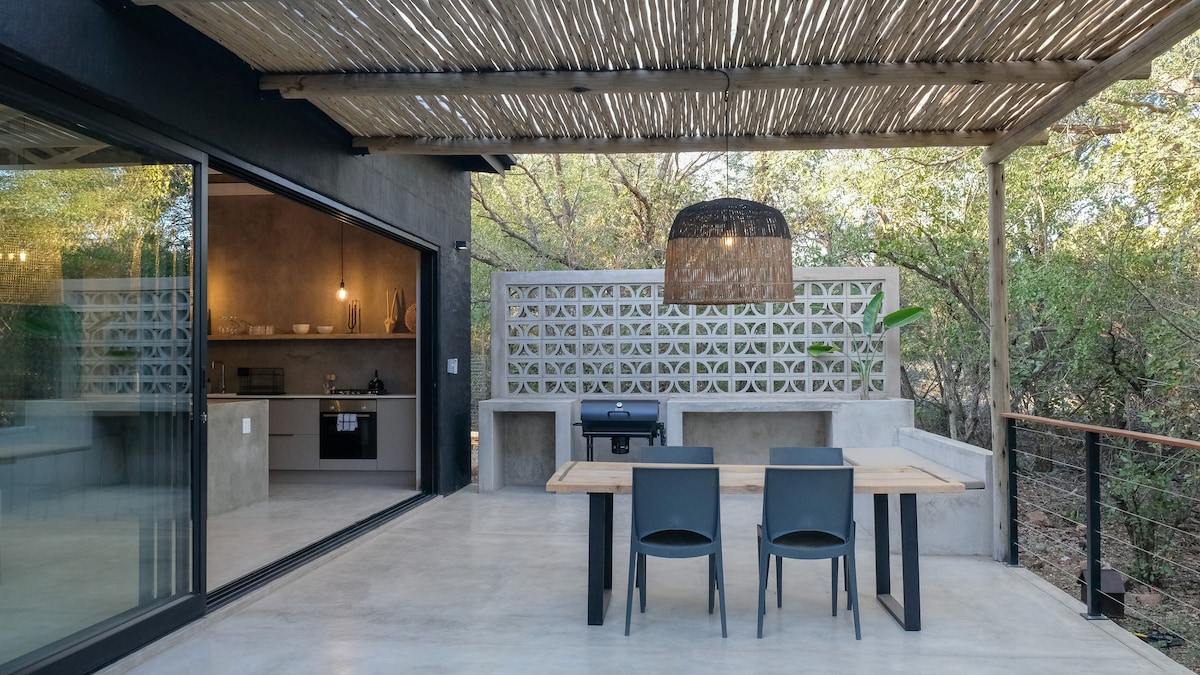 A spacious outdoor terrace features a dining table with two chairs beneath a shaded overhang. A grilling area is included, alongside a minimalist design that incorporates natural elements, allowing for views of the surrounding bush landscape.