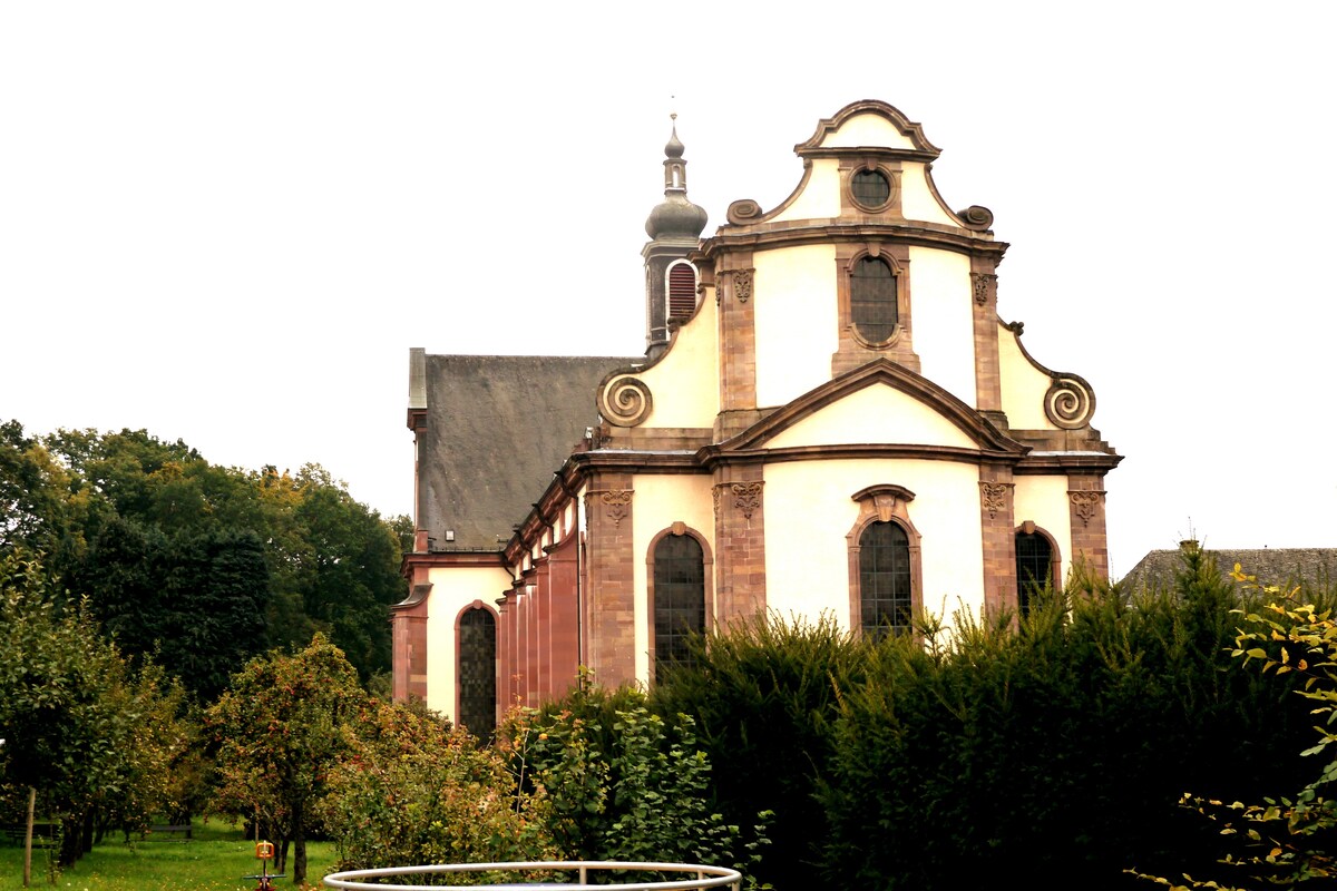 The image depicts a historic building characterized by its distinct architectural features, including symmetrical windows and a prominent bell tower. Lush greenery surrounds the structure, adding to the serene atmosphere of the location.
