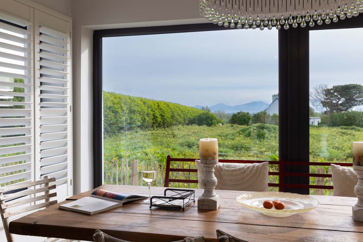 A dining area features a rustic wooden table surrounded by elegant chairs. A large window showcases a view of lush greenery and distant hills, while soft light filters through, creating a calm atmosphere. Decorative candles and a fruit bowl are present on the table.