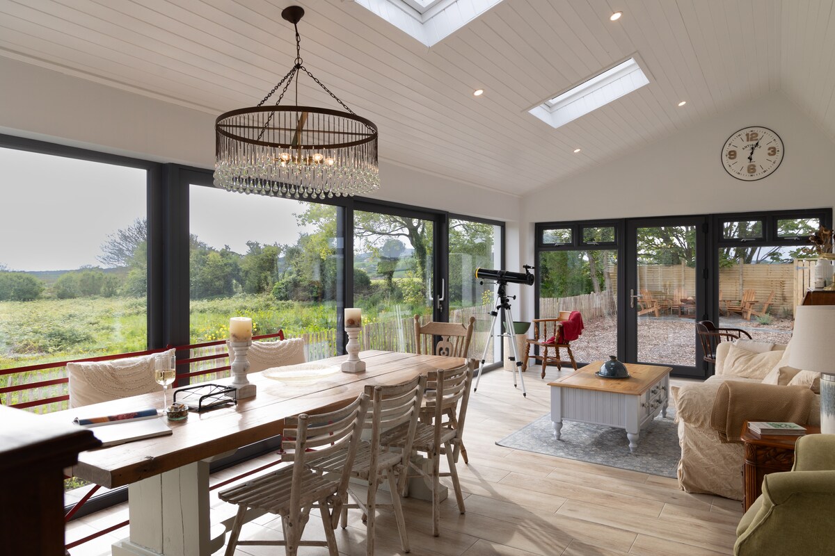 A spacious living area features a large dining table surrounded by wooden chairs. Tall glass doors provide views of the greenery beyond. A telescope is positioned near the window, while a chandelier hangs gracefully from the ceiling, illuminating the room with soft light.