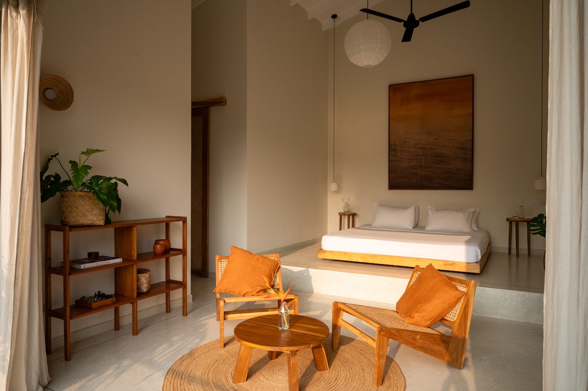 An airy room is filled with natural light, featuring a spacious bed and a cozy seating area. Two wooden chairs with orange cushions are positioned around a small table. A plant and decorative items add warmth, while a large piece of art enhances the subtle color palette.