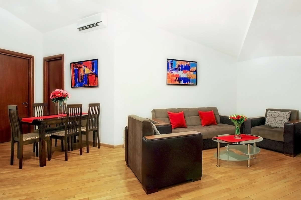 A living space features a cozy sofa set and a round glass coffee table, surrounded by art pieces on the walls. A dining area with a wooden table and chairs is visible, complemented by a vase of flowers. Natural light enhances the warm wooden flooring.