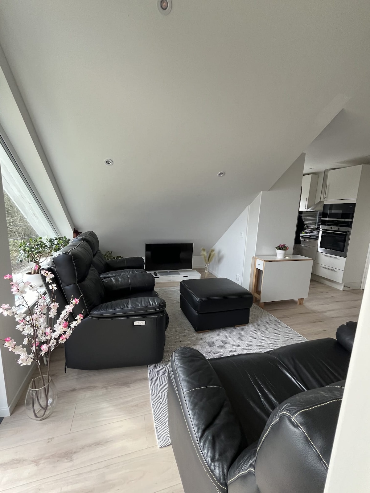 A modern living area is furnished with black leather seating, including a pair of recliners and an ottoman. A small table sits nearby. A flat-screen television is positioned against the wall, with light streaming in through large windows that offer a view of the surroundings.