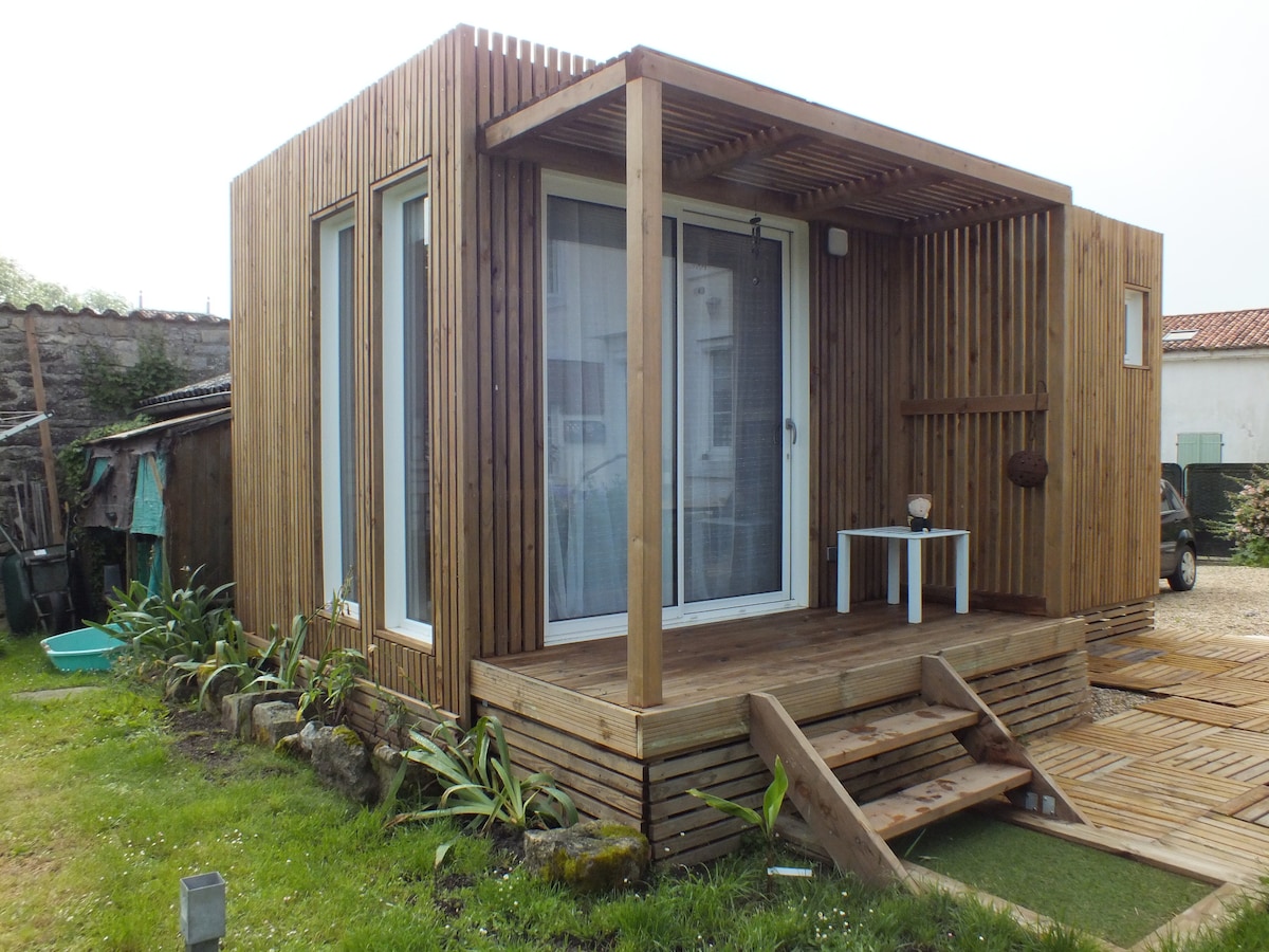 A wooden cabin features a structured facade with vertical slats, large glass doors, and a covered porch area. Steps lead up to a small outdoor space adorned with plants along the sides, providing a seamless blend of nature and comfort.