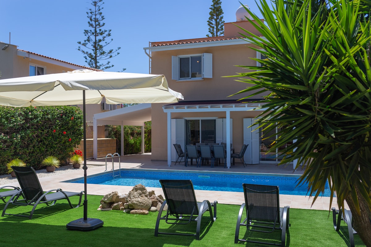 A well-maintained outdoor area features a swimming pool bordered by green grass. Lounge chairs are arranged around the pool, and a shaded dining area with a large table is visible under a canopy. Lush plants and trees surround the space, enhancing the tranquil setting.