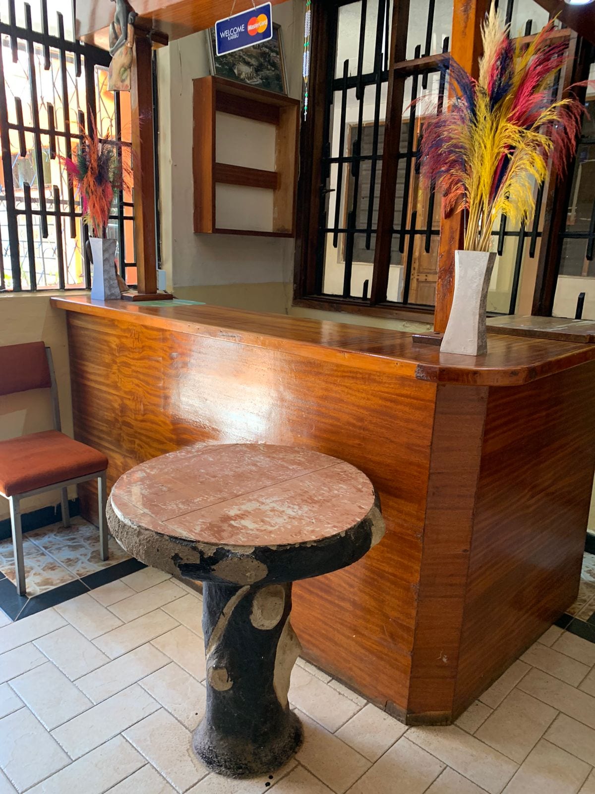 A reception area is displayed with a polished wooden counter featuring decorative vases with colorful dried flowers. A round stone table is positioned nearby, complemented by two upholstered chairs in warm tones. Large windows allow natural light to brighten the space.