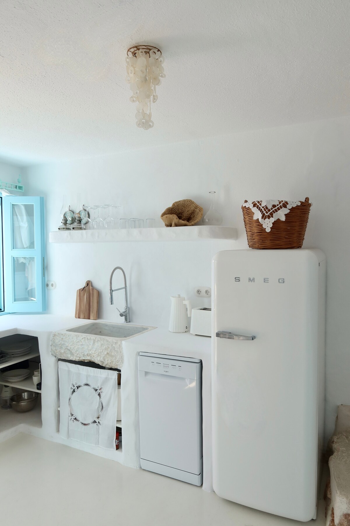 A bright kitchen area features a classic SMEG refrigerator and a modern washing machine, both set against white walls. Shelving displays glassware, with a woven basket adding a natural element. Light filters in through a blue-framed window, enhancing the clean and airy atmosphere.