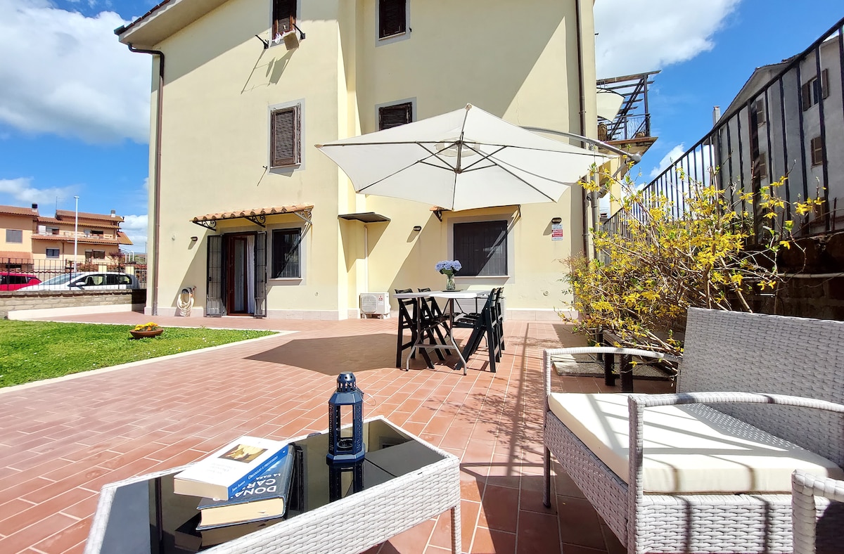 An outdoor patio area features a large umbrella providing shade over a dining table with six chairs. A cozy seating area includes two armchairs and a coffee table, creating a welcoming space for relaxation. Lush greenery is visible in the background.