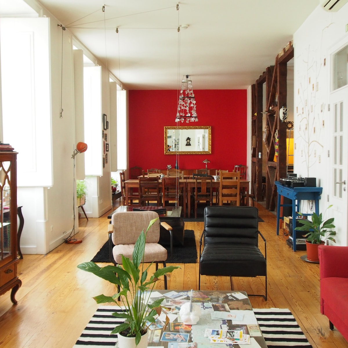 A spacious common area features a mix of comfortable seating, including a black armchair and a beige chair. A dining table with wooden chairs sits under ample natural light from large windows. A vibrant red wall is complemented by a decorative mirror and various plants scattered throughout.