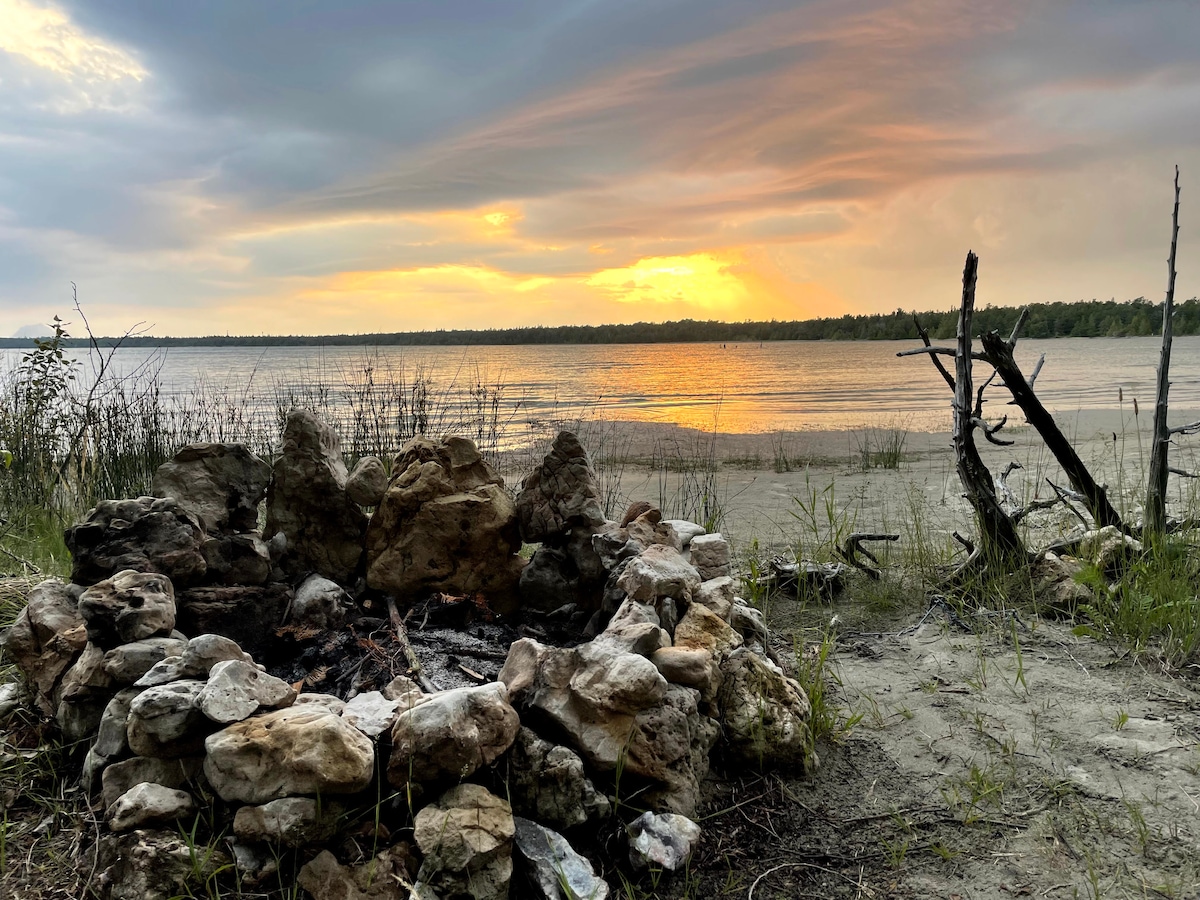 A stone fire pit is positioned on the sandy beach, surrounded by grass and sparse vegetation. The calm water reflects a warm sunset, while a shoreline extends in the background, providing a serene view of nature and the horizon.