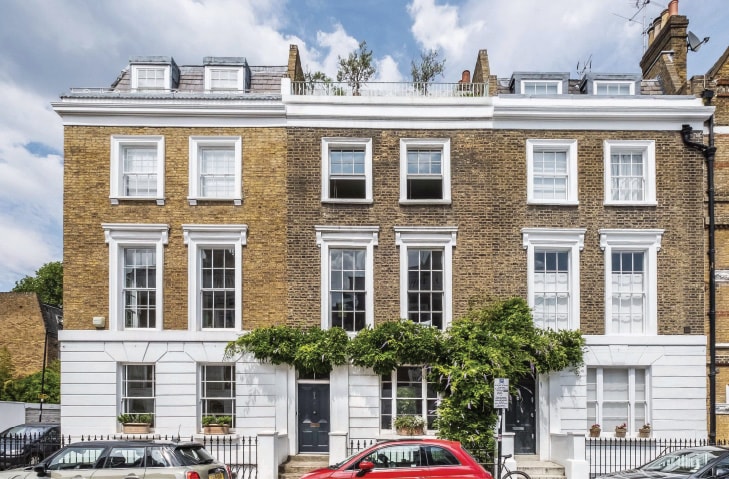A five-storey town house is presented in warm brick, complemented by white trim. Large windows enhance the façade, allowing natural light to flood the interior. Green plants adorn the entrance, and a small garden is visible at the ground level, creating a pleasing visual connection to the surroundings.