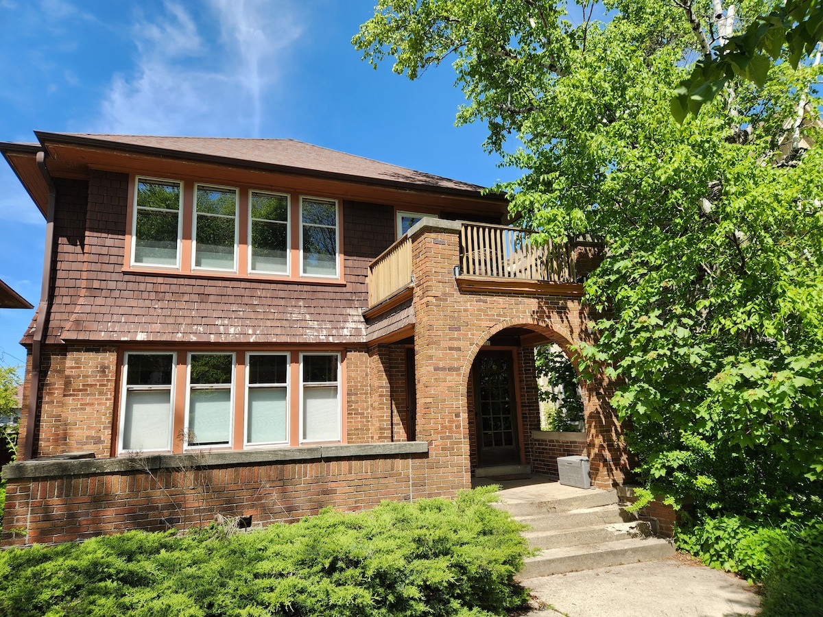 The exterior of the upper-floor apartment features a brick facade adorned with original woodwork. Large windows invite natural light, while a balcony extends from the upper level. Green foliage surrounds the entrance, providing a welcoming ambiance and a connection to nature.