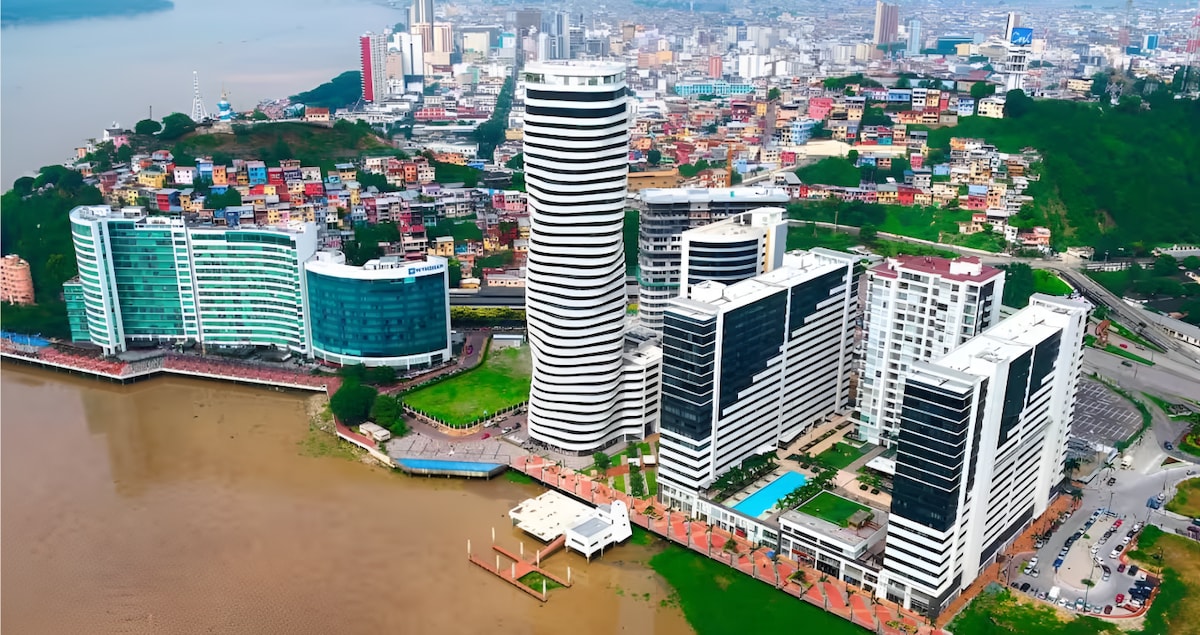 An aerial view captures the vibrant skyline of Guayaquil, showcasing modern high-rise buildings alongside colorful homes. The waterfront is visible, with a pool area and walkways highlighted. The lush greenery on the hills provides a natural backdrop to the urban landscape.