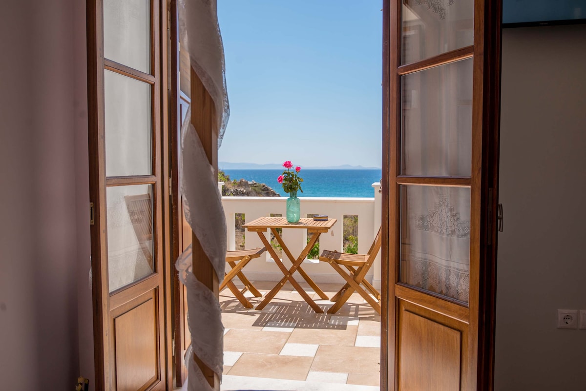 A balcony view is presented through open wooden doors, revealing a small wooden table with a vase of pink flowers. The sea glimmers in the background, showcasing shades of blue under clear skies.