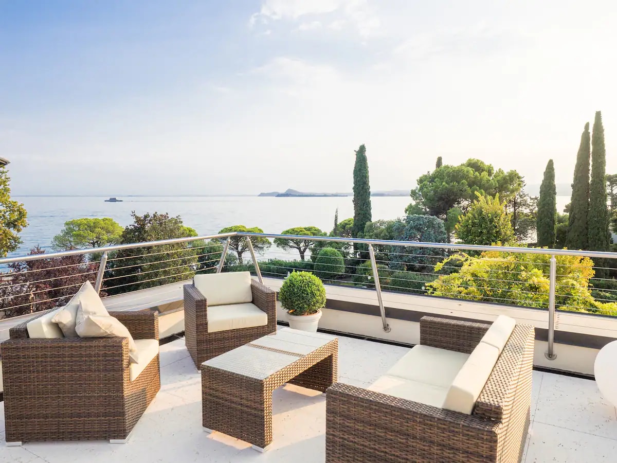 An outdoor balcony features comfortable seating arrangements with wicker furniture, including a coffee table and multiple chairs. Lush greenery and trees line the space, offering views of the expansive ocean under a clear sky.