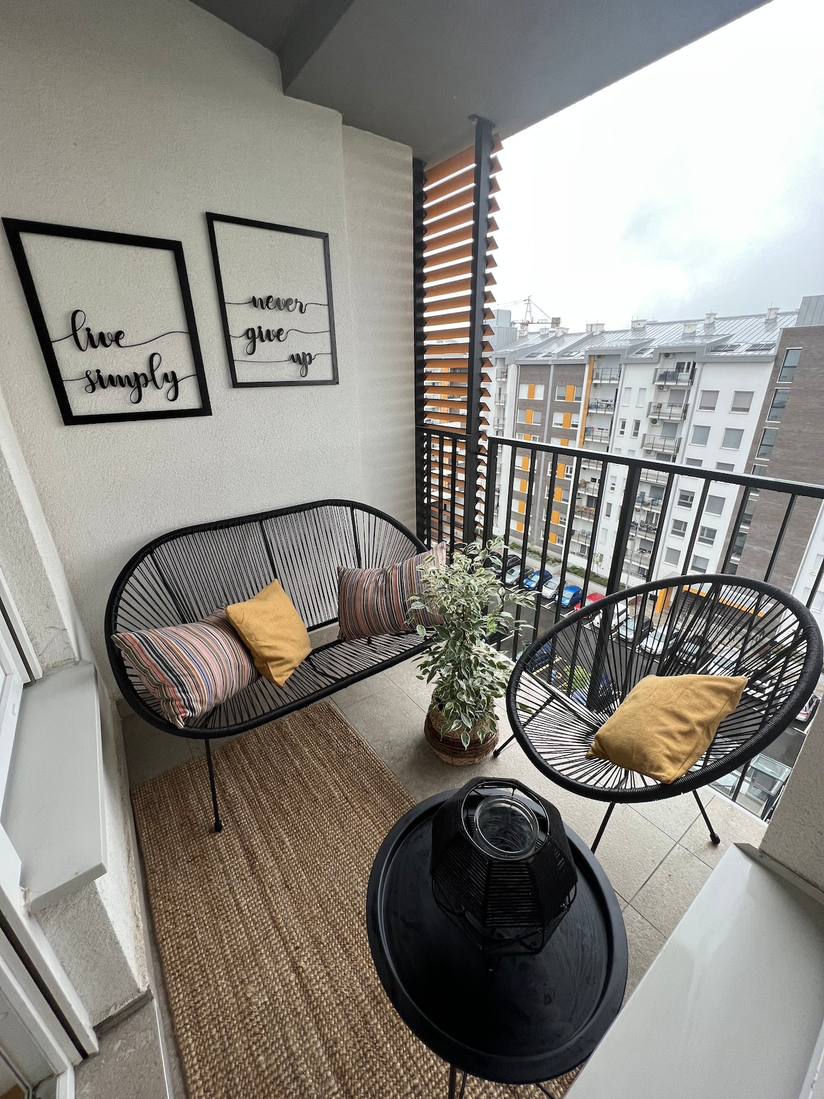 A balcony space is presented with a comfortable two-seat sofa adorned with patterned cushions. An additional chair accompanies a small round table. Two framed prints with motivational phrases are hung on the wall, complemented by a potted plant. Light filters through partially closed slats.