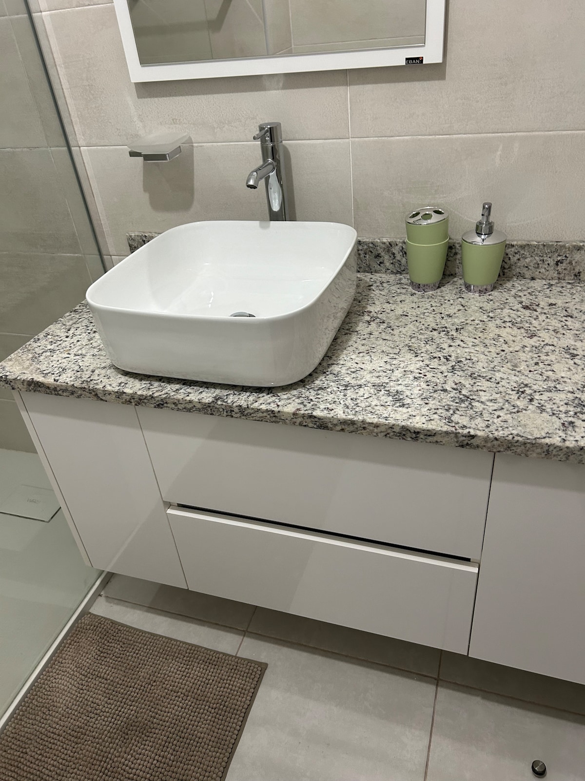 A clean bathroom countertop is featured, showcasing a white square sink atop a smooth granite surface. Two green cups and a dispenser sit next to the sink, complementing the modern cabinetry below. The glass shower enclosure stands nearby, creating a sleek appearance.