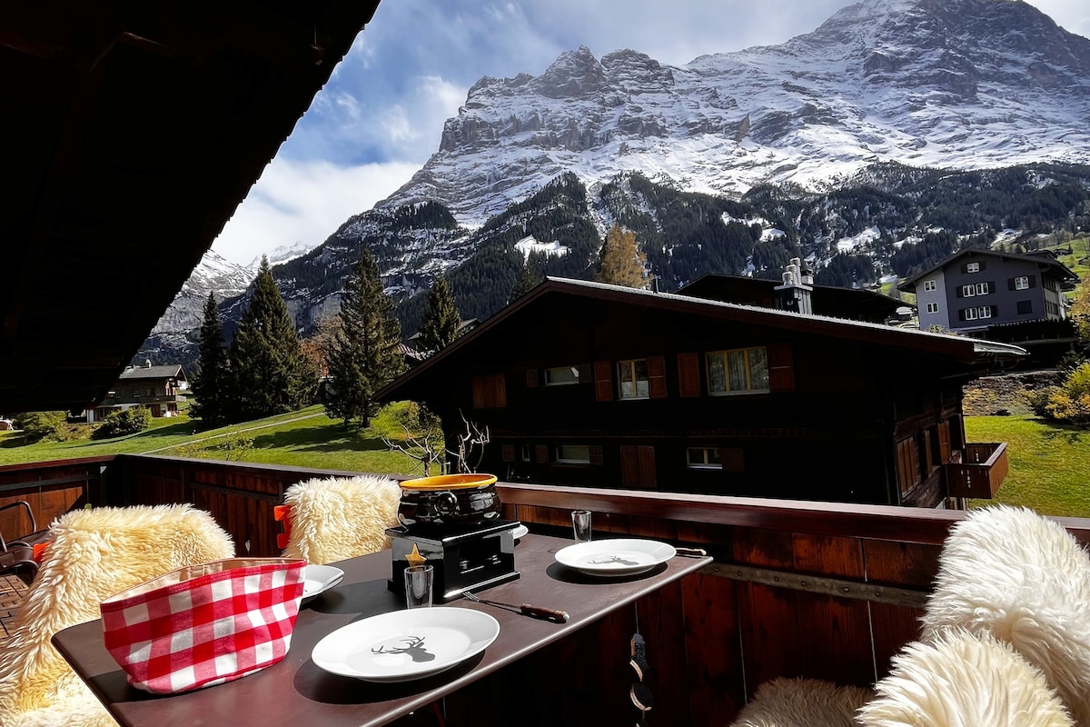 A balcony area features a rustic dining table set with plates and a grill, offering a view of snow-capped mountains and lush greenery. Soft, plush throws adorn the chairs, enhancing comfort in the outdoor space.