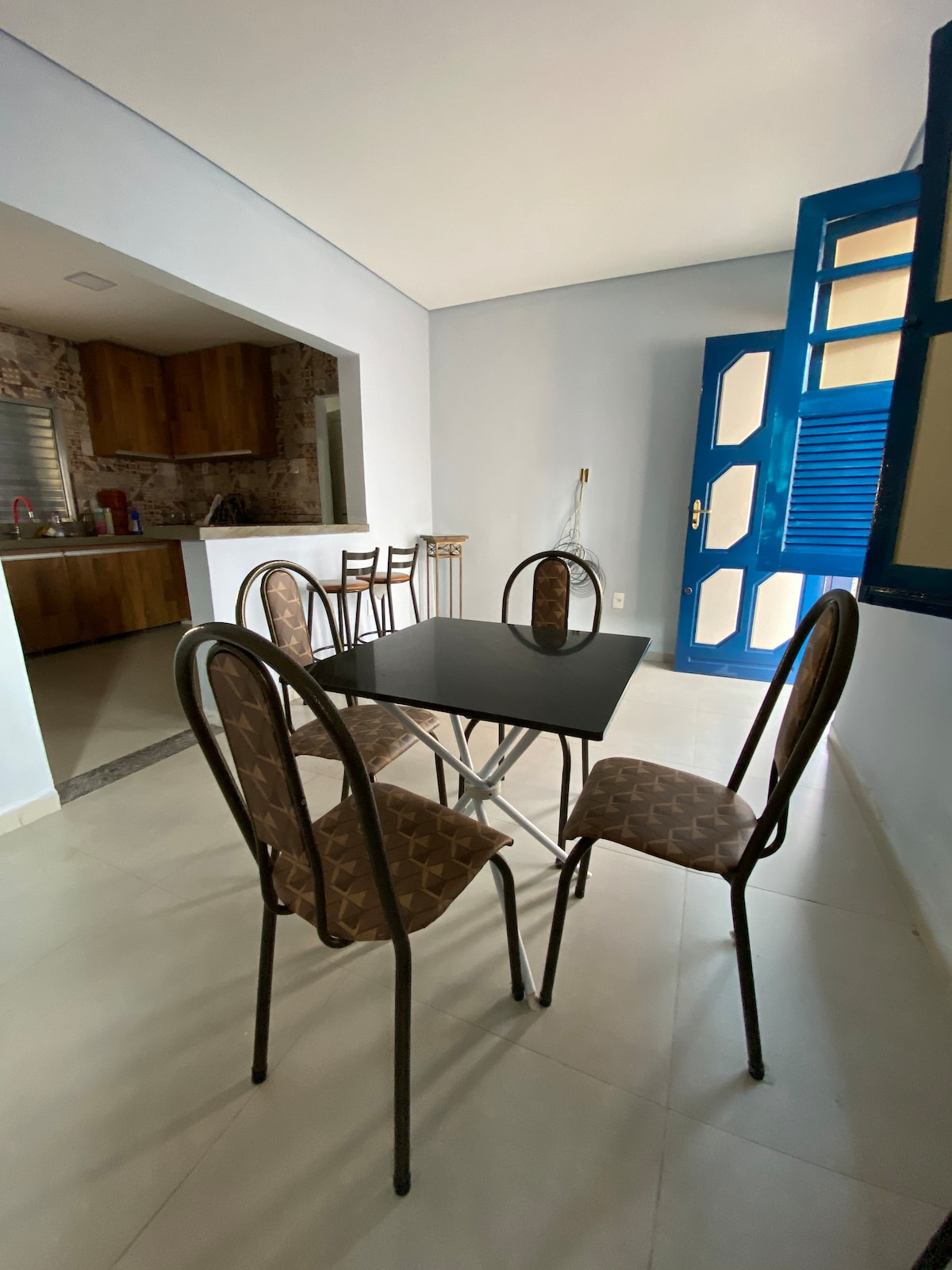A dining area features a square black table surrounded by four patterned chairs. Natural light enters through a nearby blue window, illuminating the space. An open doorway leads to the kitchen area, which is partially visible in the background.
