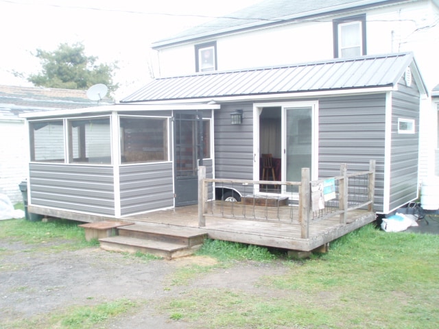 An exterior view of a two-story building showcasing a wooden deck with a railing. A screened-in porch extends from the main structure, while the main entrance is visible through glass doors. A small lawn area surrounds the property, contributing to the rural setting.