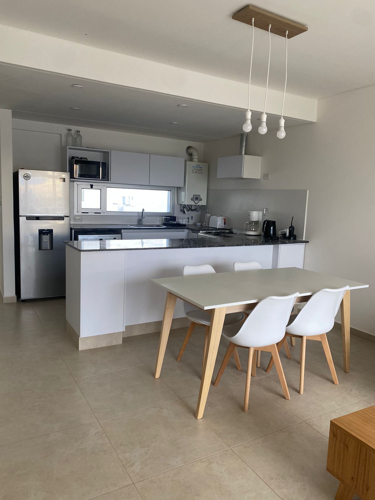 A bright dining area features a light wood table with three white plastic chairs. Modern kitchen appliances are visible in the background, along with a sleek refrigerator. Soft natural light illuminates the space, highlighting the clean lines and open layout.