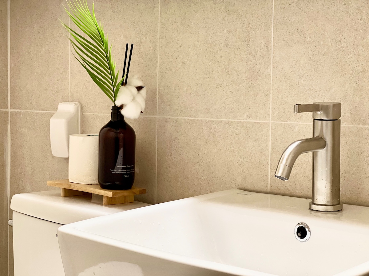 A modern bathroom sink is featured, accompanied by a stylish liquid soap dispenser and a small vase containing decorative greenery and cotton. Soft lighting highlights the sleek surfaces and neutral tile backdrop, creating a clean and welcoming atmosphere.