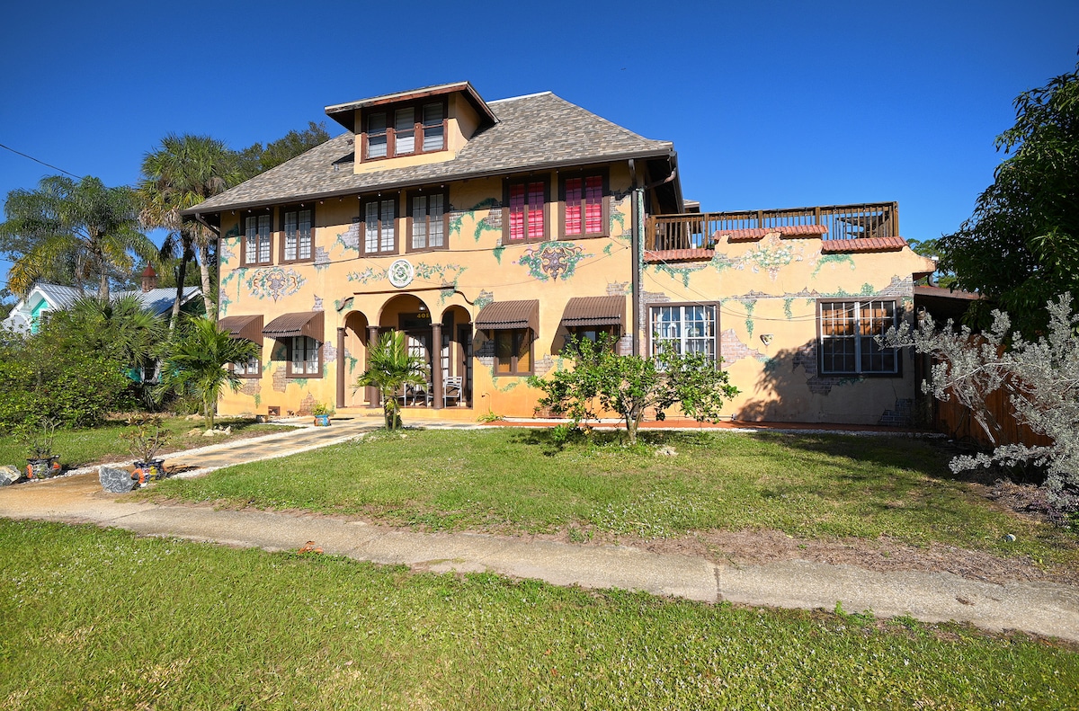 A charming exterior of Casa Coquina Del Mar showcases a two-story building adorned with colorful murals. Lush greenery surrounds the pathways, leading to the entrance. The second-floor deck offers a space to relax, while numerous windows and shutters add character to the structure.