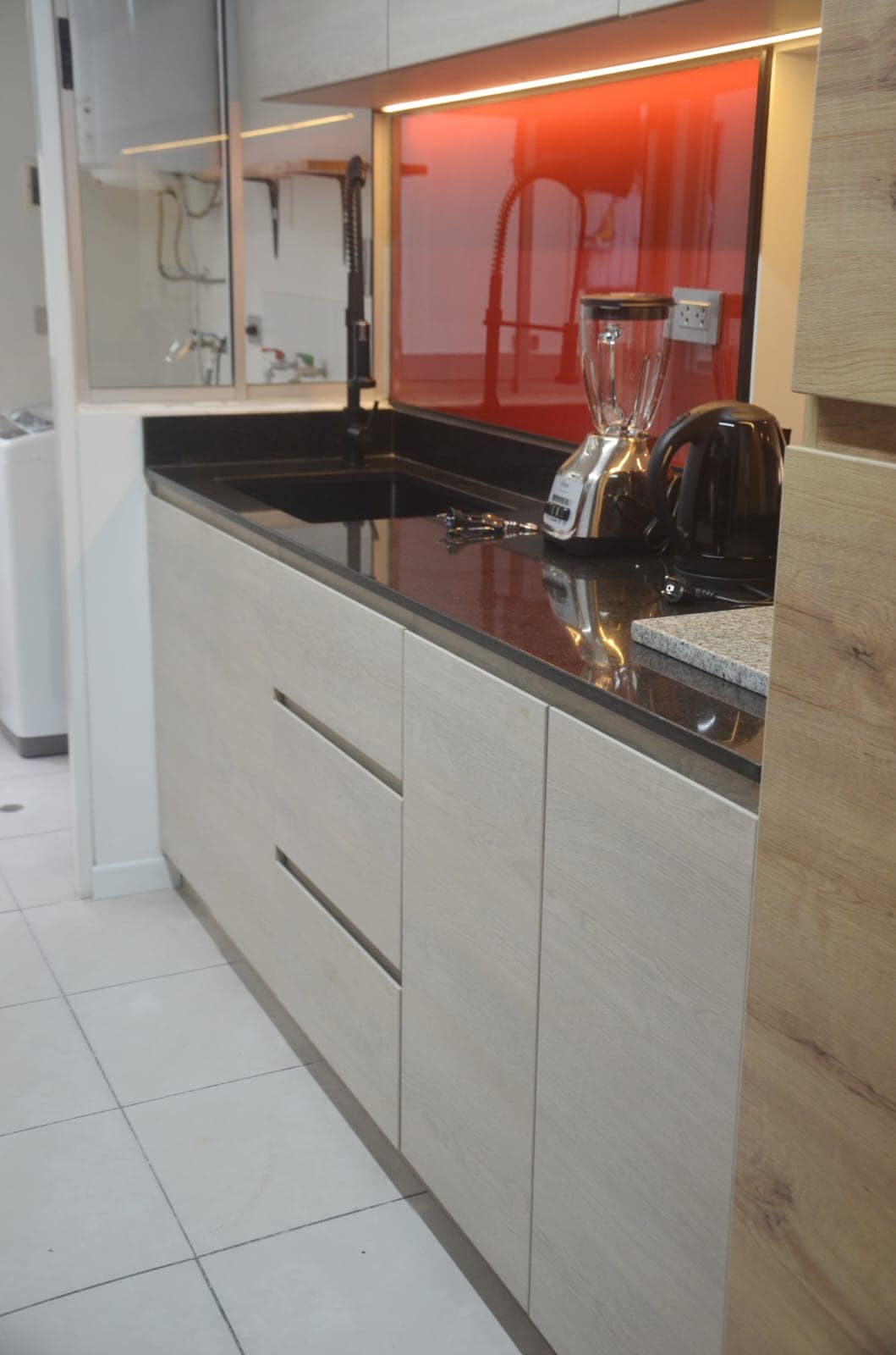 A modern kitchen area is visible, featuring a black countertop and light wood cabinetry. A blender and electric kettle are placed on the counter. A red backsplash adds a splash of color, while warm lighting illuminates the workspace.