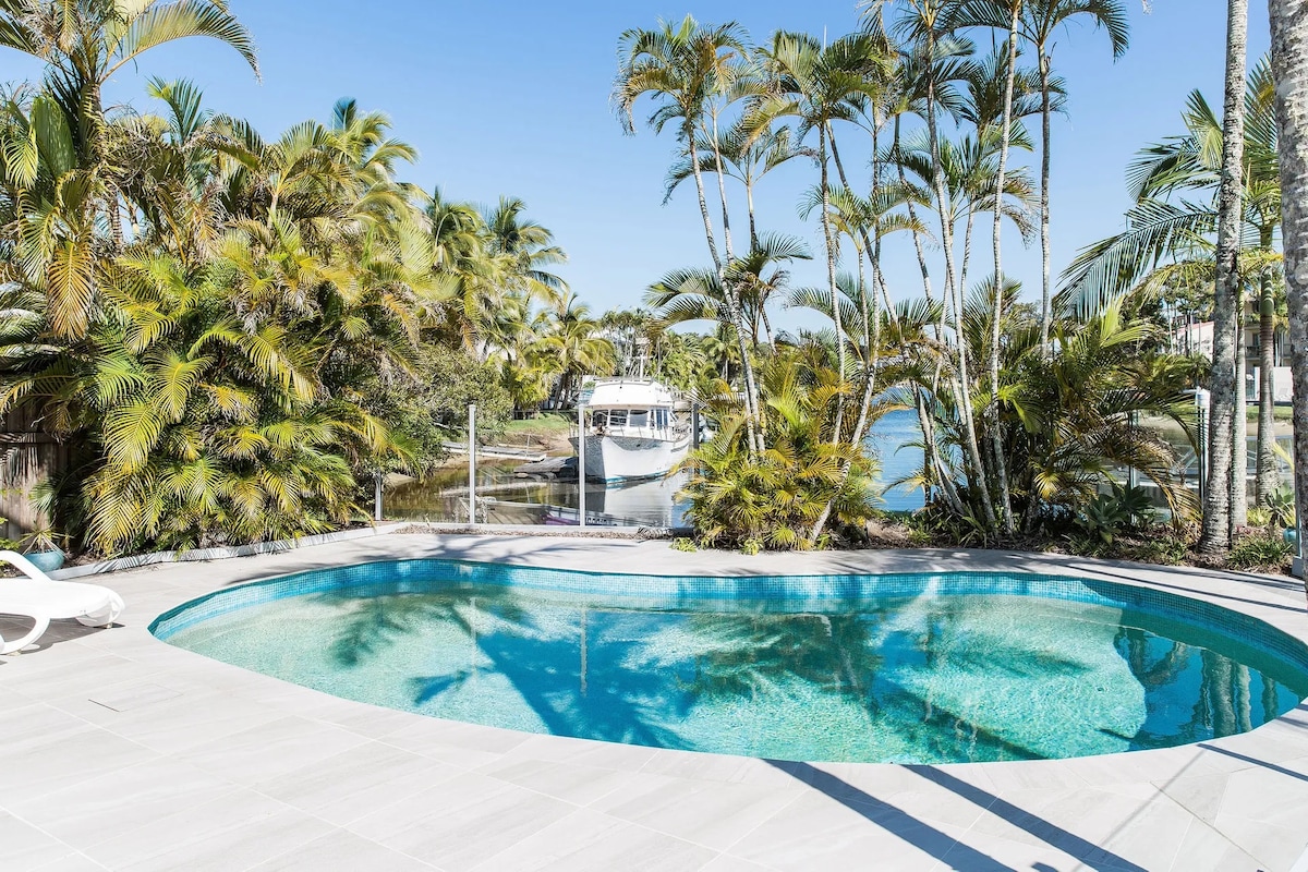 A serene lagoon-style pool is surrounded by lush palm trees, offering a tranquil setting. The water reflects the clear blue sky, with a boat visible in the distance, highlighting the proximity to water activities.