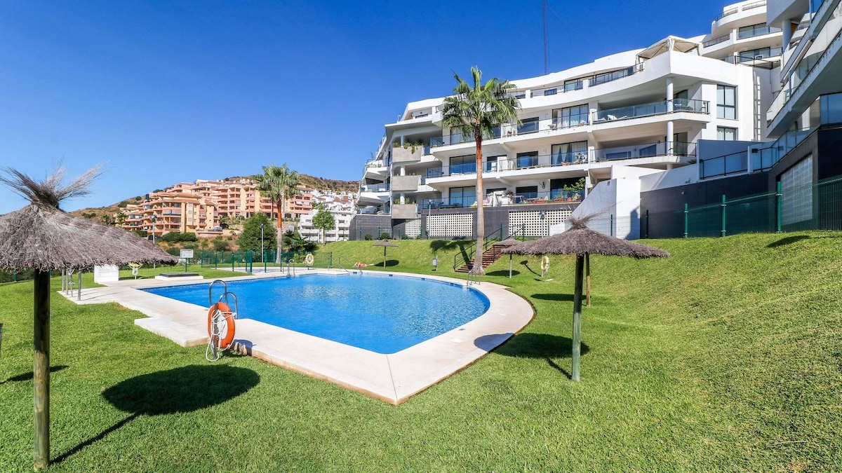 A landscaped area features a large swimming pool surrounded by lush green grass. Thatched umbrellas provide shade, and a lifebuoy is mounted next to the pool. Modern apartment buildings are visible in the background, highlighting the inviting outdoor space.