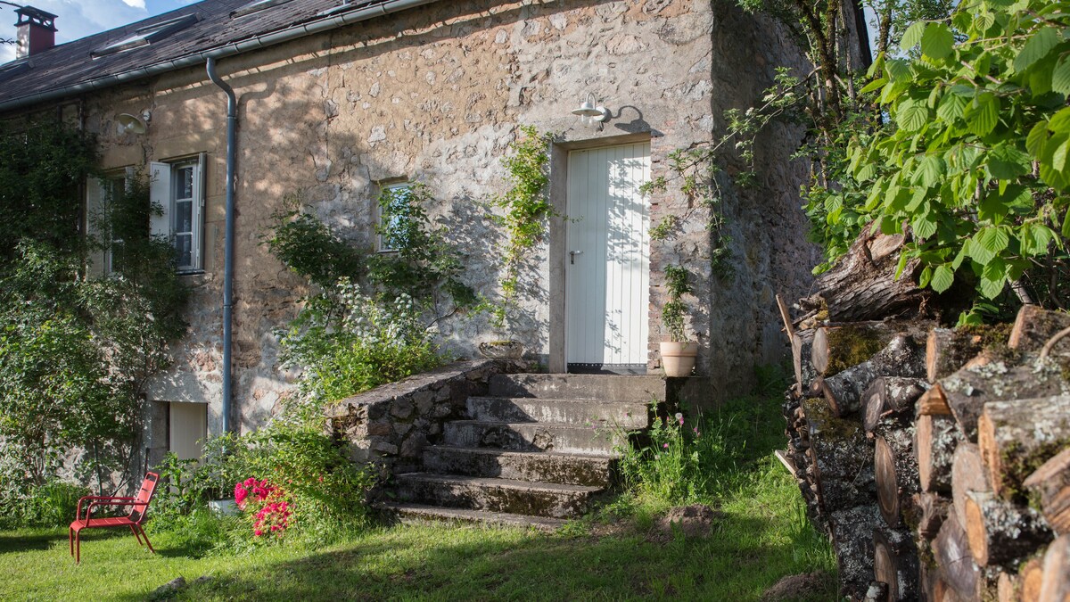 An entrance to a stone house is framed by lush greenery and outdoor steps. A potted plant sits near the doorway, while a stack of firewood is positioned to the side. A single red chair adds a touch of color to the serene outdoor setting.