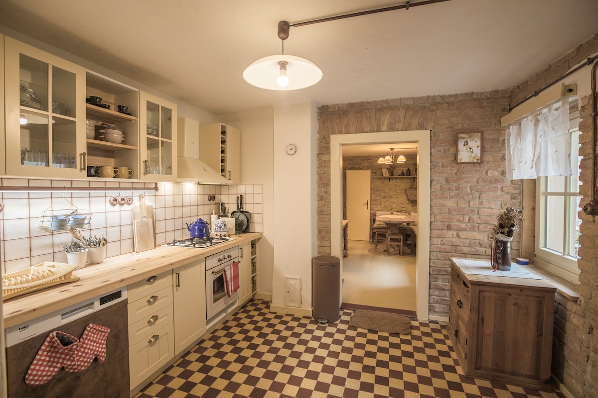 A fully equipped kitchen features wooden countertops and cream-colored cabinets. The checkered floor adds character, while various kitchen utensils and a blue kettle are arranged on the countertop. A doorway leads to a dining area, creating a connected space for meal preparation and enjoyment.