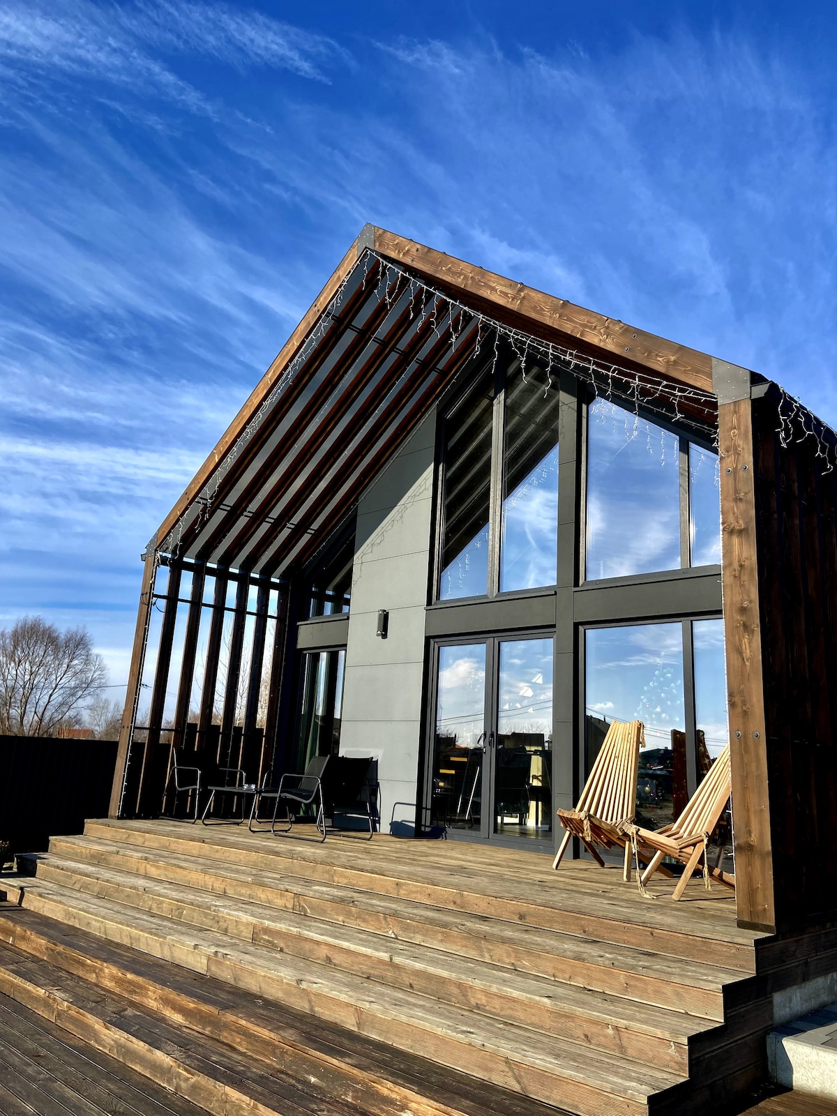 A modern cabin features a steeply pitched roof and large glass windows, allowing natural light to fill the interior. A spacious deck with wooden steps leads to seating areas, including two striped lounge chairs, perfect for outdoor relaxation.