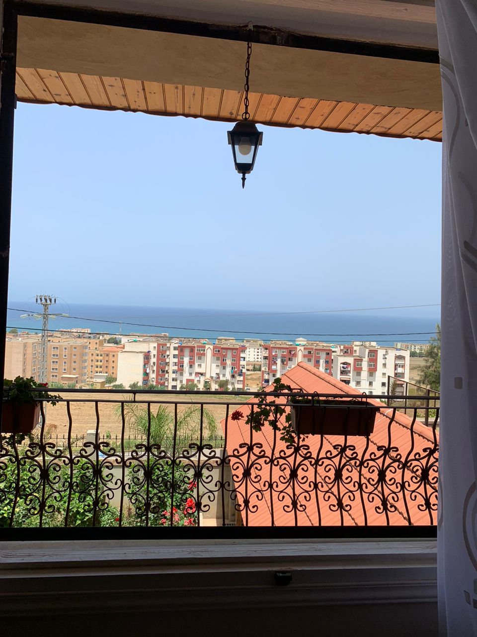 A view from a window showcases the coastline and the sea beyond, framed by a decorative iron balcony railing. Colorful houses are visible in the foreground, set against a clear blue sky.