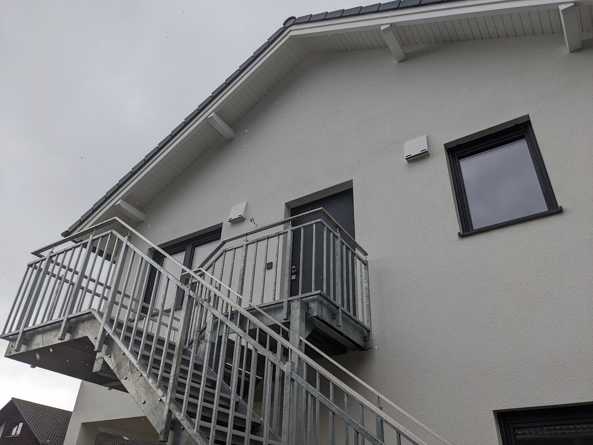 The exterior of a two-story building is shown, featuring a metal staircase leading to the upper level. Large windows are visible, framed by sleek black surrounds. The light-colored exterior walls reflect a modern architectural style.