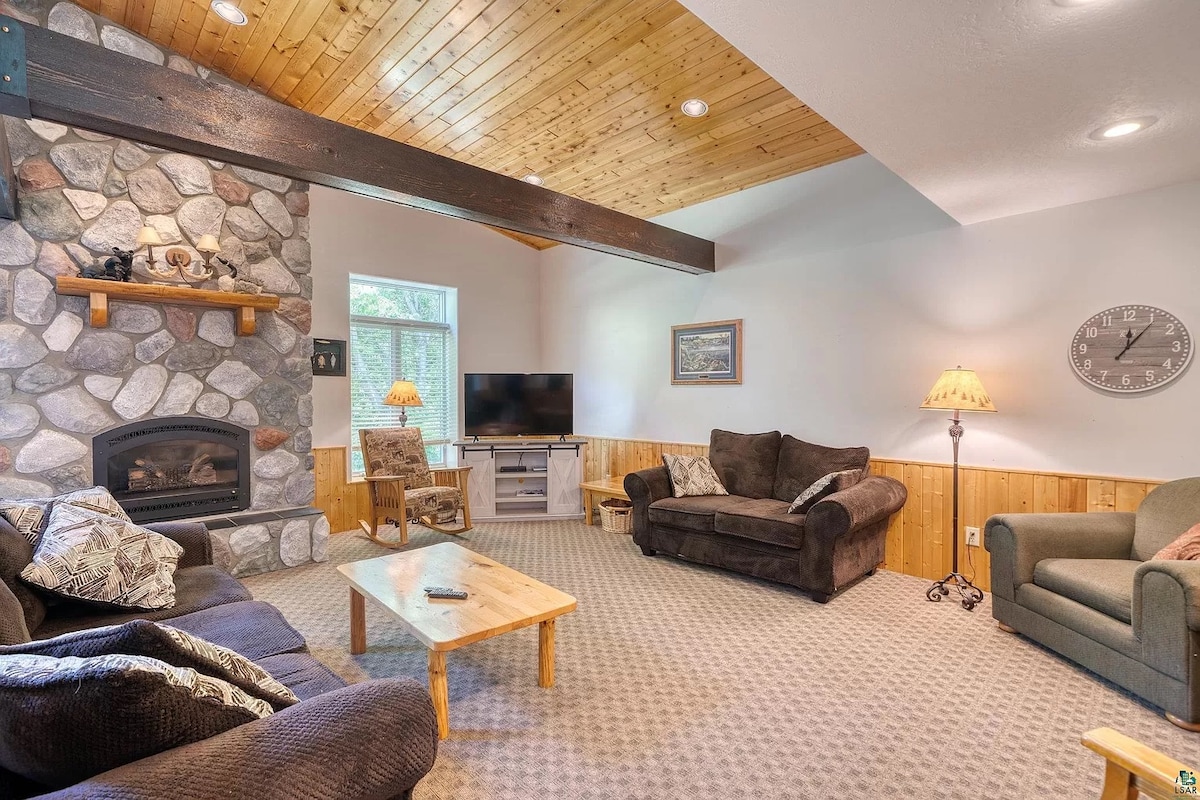 A living area features a grand stone gas fireplace as a central element. A wooden ceiling with exposed beams enhances the space, which includes a large seating arrangement of plush couches and armchairs. Natural light enters through a window, illuminating the cozy atmosphere.