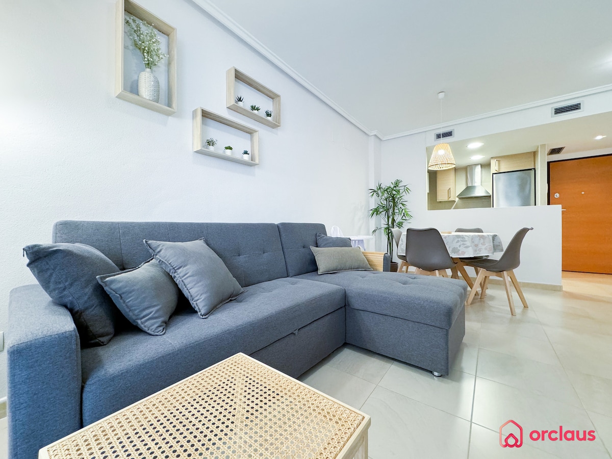 A modern living space features a gray sectional sofa adorned with matching throw pillows. A wooden coffee table with a textured surface is positioned in front of the seating area. In the background, a dining area includes a round table surrounded by sleek chairs, with a glimpse of the kitchen visible.