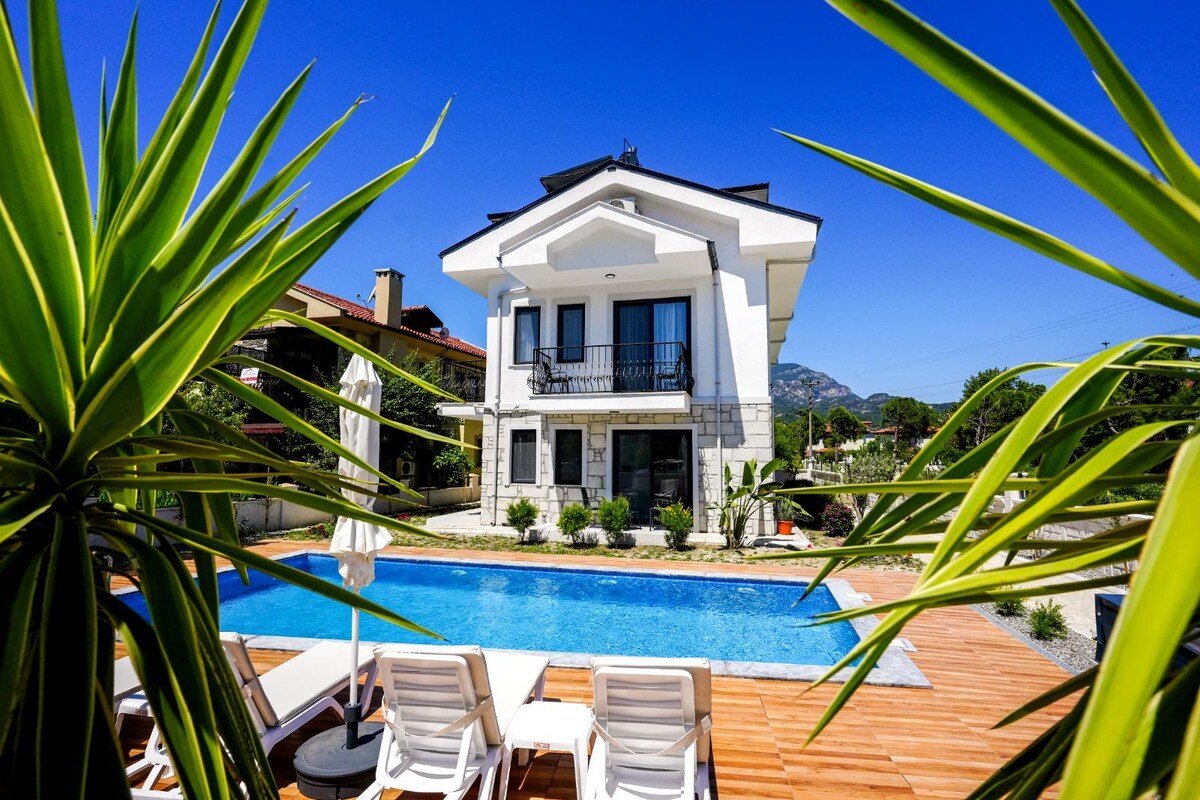 A modern two-story house features large glass doors that open to a serene outdoor area. The backyard presents a clear pool surrounded by lounge chairs and palm plants, with vibrant greenery enhancing the peaceful setting under a bright blue sky.