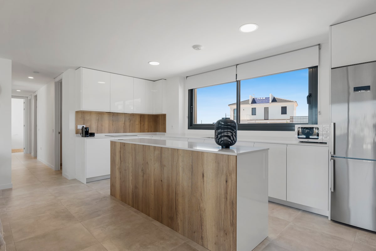 A modern kitchen space features sleek white cabinetry paired with a natural wood accent on the island. A large window allows natural light to brighten the area, while appliances such as a microwave and stainless steel refrigerator are visible.