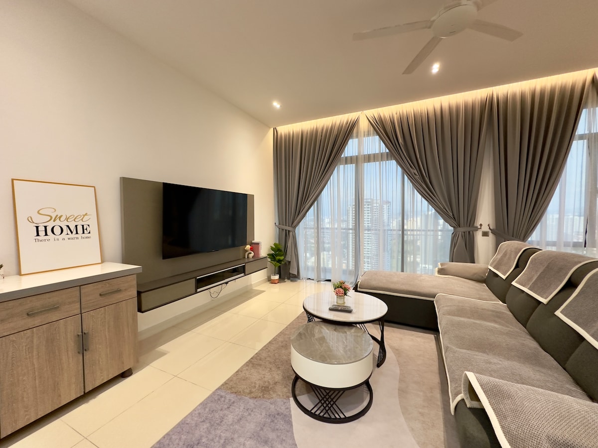 A modern living hall features a large flat-screen TV mounted on the wall. A comfortable L-shaped sofa is arranged next to a coffee table, with decorative plants and a sign reading 'Sweet HOME' positioned on a cabinet. The space is brightened by natural light from large windows dressed with flowing curtains.