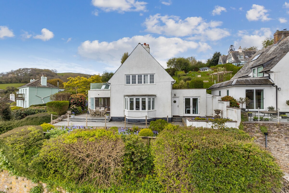 The detached cottage is set on an elevated position surrounded by lush gardens and greenery. A spacious outdoor deck offers seating for relaxation, with scenic views of the countryside and nearby homes. The white exterior is complemented by large windows, inviting natural light into the interiors.