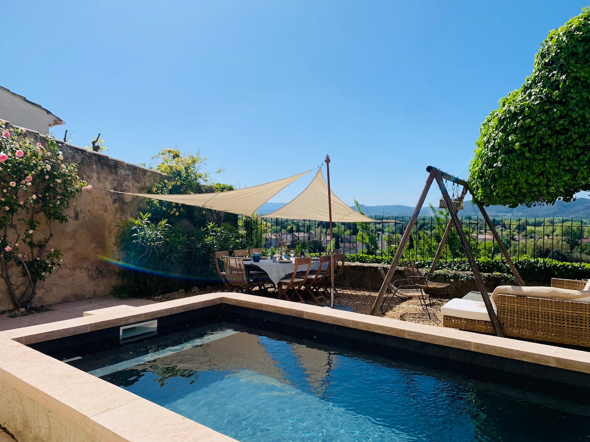 A serene outdoor space features a refreshing swimming pool surrounded by natural stone. A large shade sail provides shelter over a dining area with a wooden table and chairs. Lush greenery and a swing enhance the inviting ambiance, with views of the surrounding landscape visible beyond.