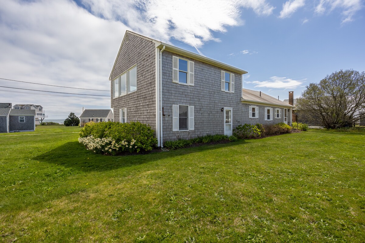 A spacious two-story home is surrounded by a lush green lawn and vibrant shrubbery. The exterior features light grey shingles and large windows, allowing natural light to brighten the interior. The setting is complemented by a clear blue sky with soft clouds.