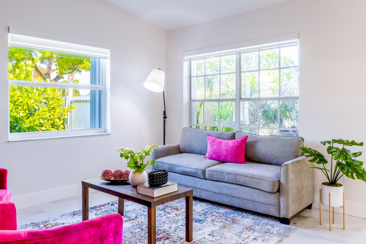 A bright living area features a light gray sofa adorned with a pink accent pillow, paired with a wooden coffee table. Two large windows allow natural light to fill the space, highlighting a colorful area rug and potted greenery.