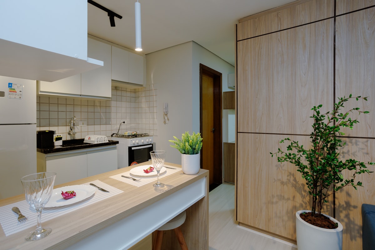 A modern kitchen area is visible, featuring a complete set of appliances, including a refrigerator and gas stove. An inviting dining table is set for two, accompanied by a decorative plant in a pot. Light wood cabinetry and neutral tones create a harmonious aesthetic.