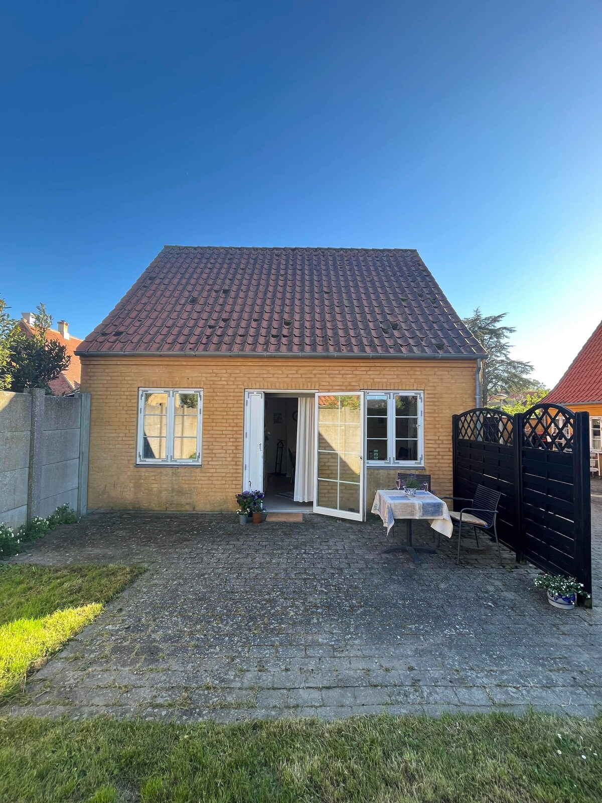 A charming house with a sloped red roof is visible, featuring large front windows and a welcoming entrance. A small outdoor terrace with a table and chairs is seen, surrounded by greenery. A black wooden gate provides access to the property.