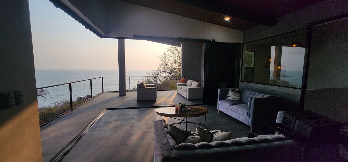 An open living area features comfortable seating arrangements that overlook a panoramic view of the ocean. Large glass doors allow natural light to fill the space, creating a bright and airy atmosphere. A round coffee table adds functionality to the seating area.