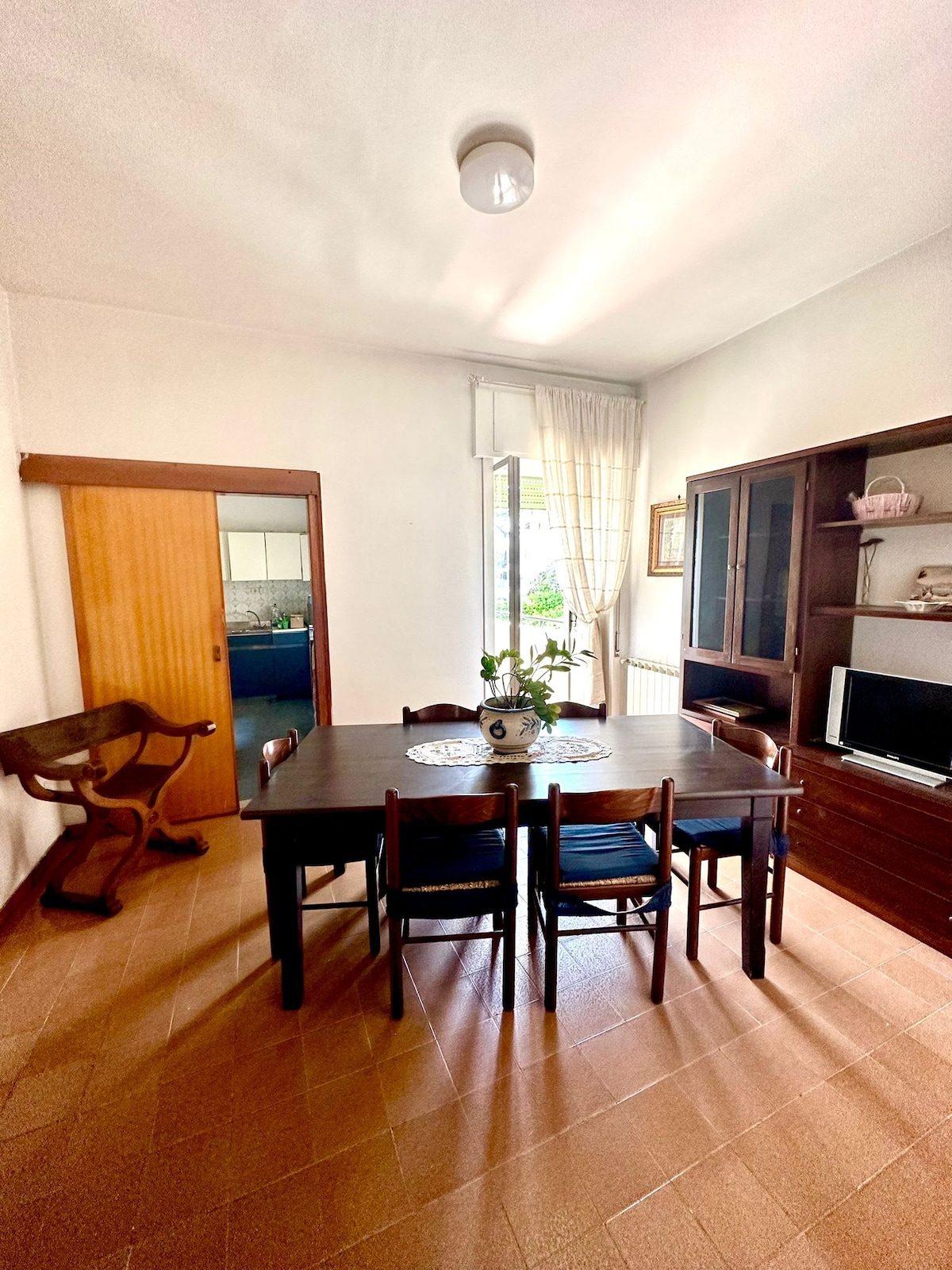 A dining area features a wooden table surrounded by six chairs, providing ample space for family meals. Natural light filters through a large window with sheer curtains, while a wooden cabinet and a small television are positioned against the wall.