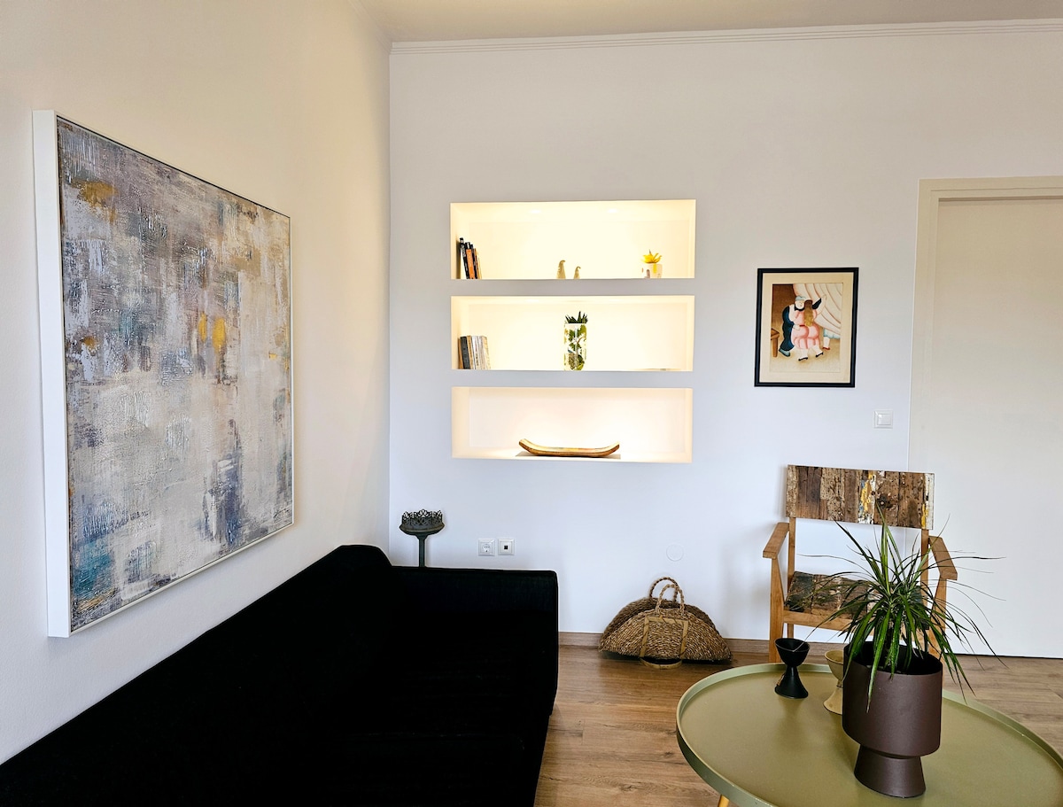 A stylish living area is observed, featuring a dark sofa against a light wall. Shelves holding art and decorative items enhance the space. A wooden chair, a plant, and a round table with a decorative centerpiece complete the atmosphere, while natural light fills the room.